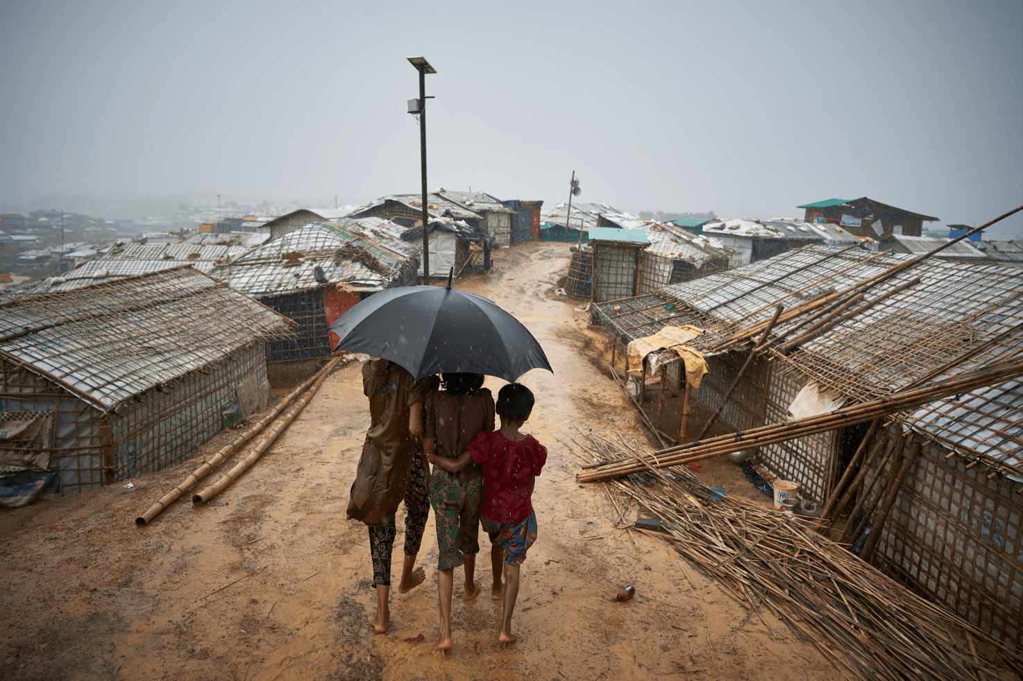 Three Rohingya children huddle under an umbrella during a downpour in Cox's Bazar, Bangladesh