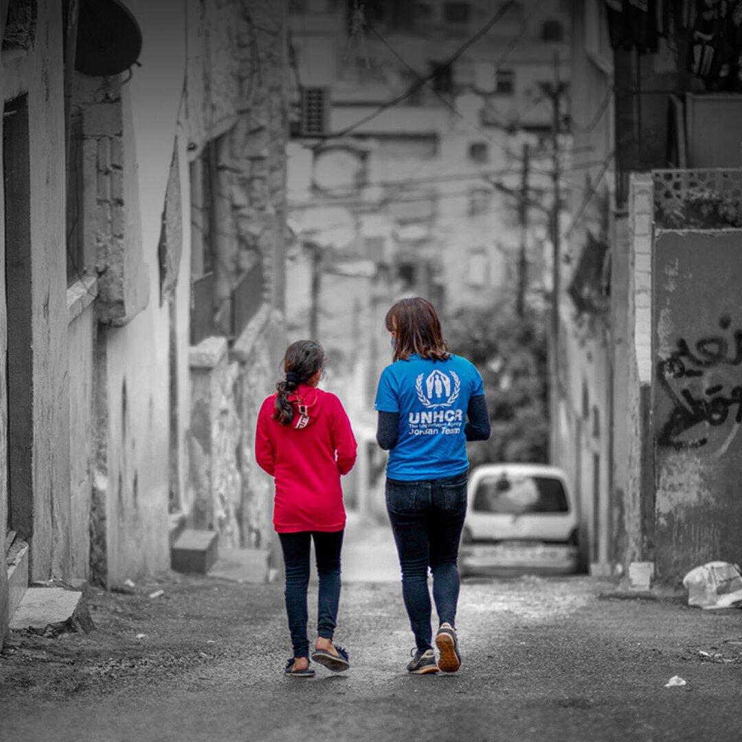 UNHCR staff and a refugee girl walking together. The background, a street seemed to be torn by war and disaster, is black and white