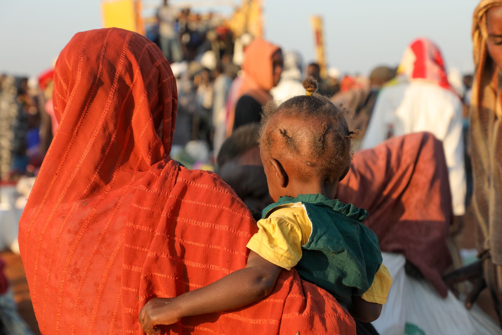 A Sudanese woman wearing an orange shawl holds her child, both with their back to the camera