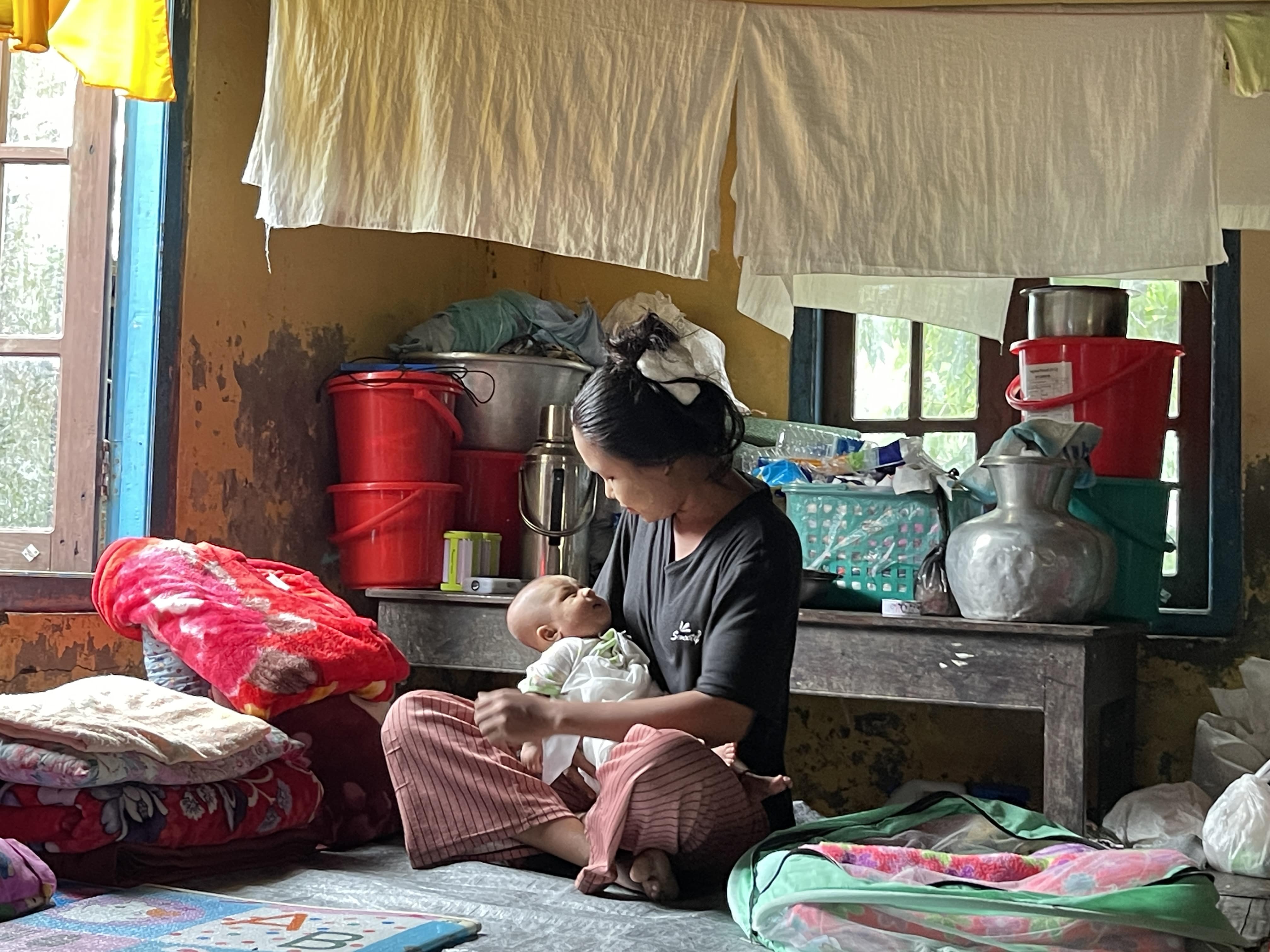 A woman sits on the floor of a simple shelter, holding a young baby. In Myanmar, humanitarian needs are only continuing to escalate.
