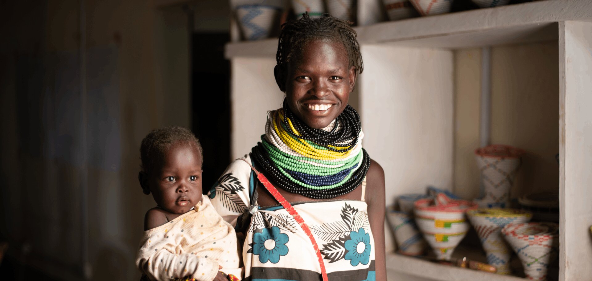 Lucy Akai, a Kenya woman, holds her baby and smiles at the camera. She is wearing brightly coloured beaded jewelery.