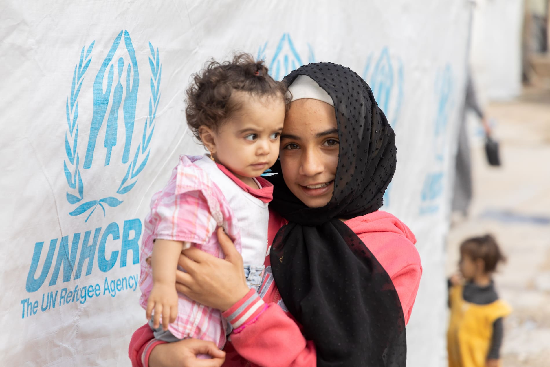 Older sister holds younger sibling outside a UNHCR emergency shelter supported by a winter kit.