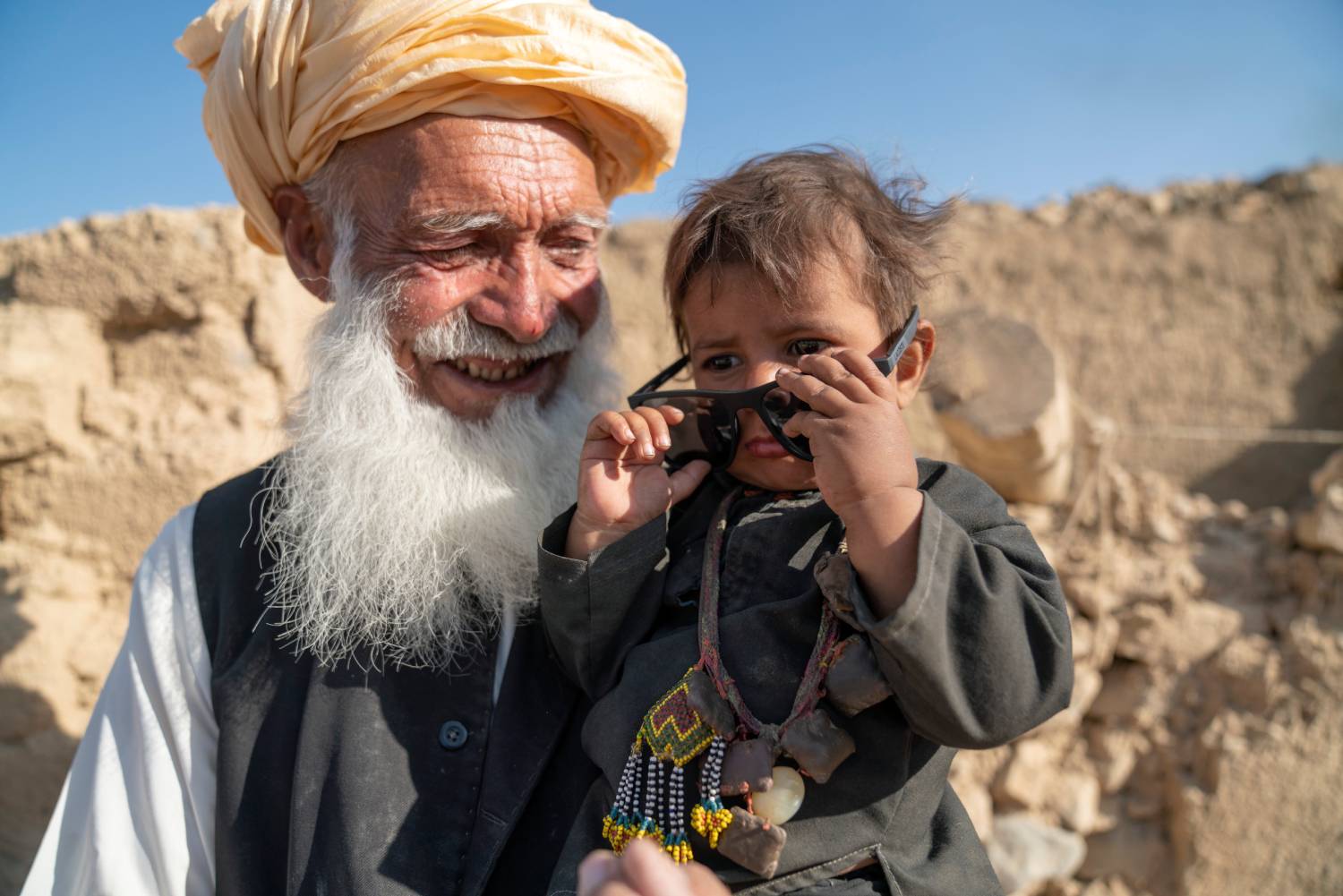 A boy puts on sunglasses while his grandfather holds him, smiling. In Afghanistan, UNHCR is providing earthquake-resilient houses for displaced families.